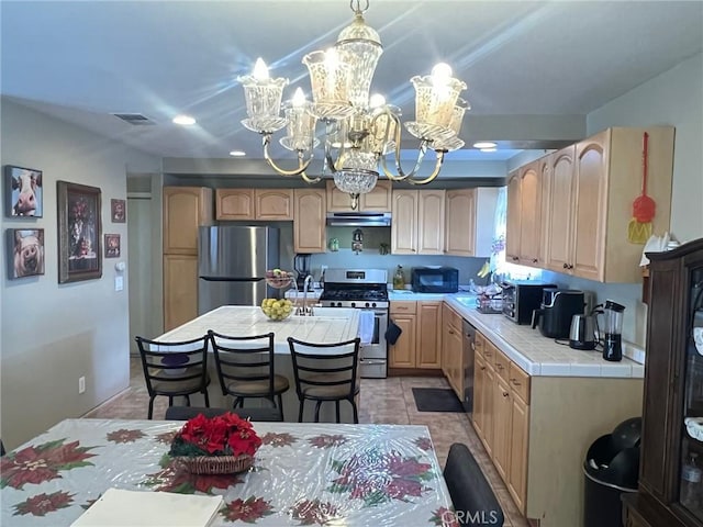 kitchen featuring appliances with stainless steel finishes, tile counters, a kitchen island, a chandelier, and light brown cabinets