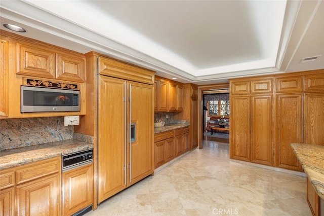 kitchen with decorative backsplash, light stone counters, a raised ceiling, and built in appliances