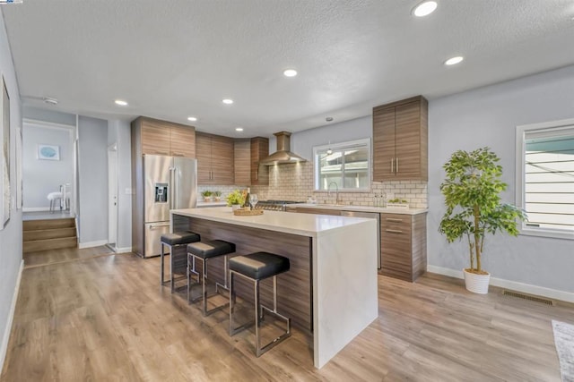 kitchen featuring tasteful backsplash, wall chimney range hood, a kitchen island, a breakfast bar, and appliances with stainless steel finishes