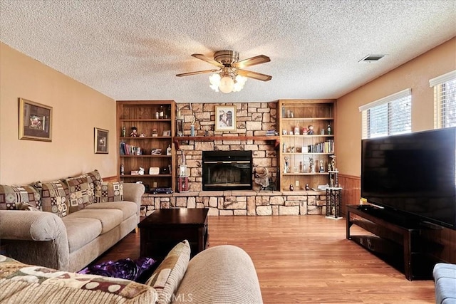 living room with a textured ceiling, built in shelves, a stone fireplace, light wood-type flooring, and ceiling fan