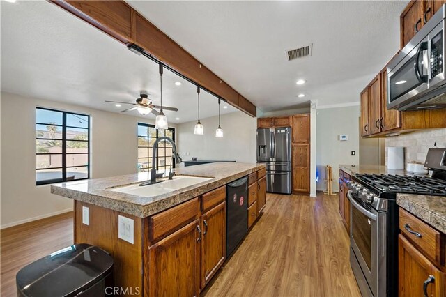 kitchen with appliances with stainless steel finishes, sink, decorative light fixtures, light wood-type flooring, and an island with sink