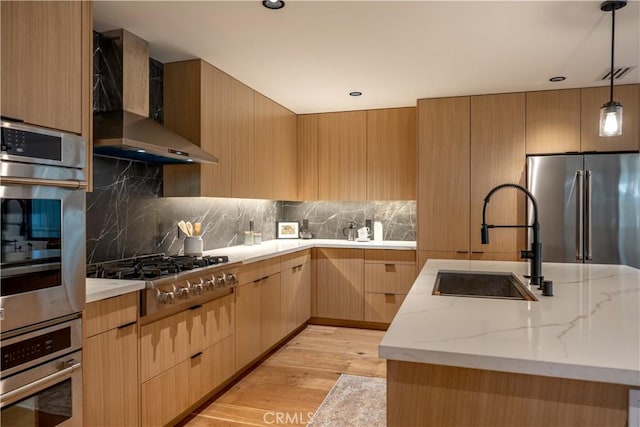 kitchen with wall chimney range hood, light wood-type flooring, light stone counters, stainless steel appliances, and a sink