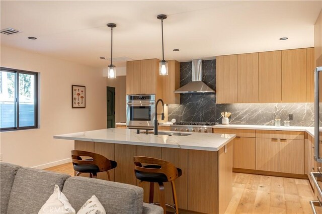 kitchen with oven, pendant lighting, a kitchen island with sink, and wall chimney range hood