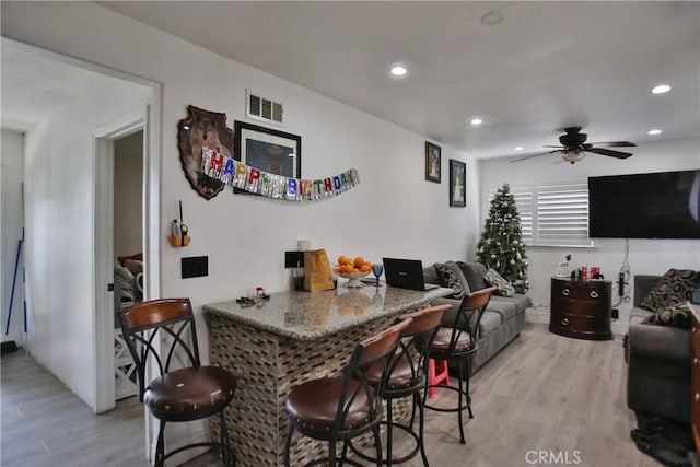 bar with ceiling fan, stone counters, and light wood-type flooring