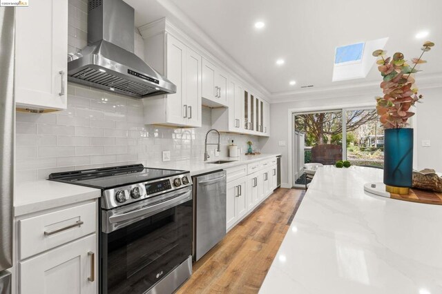 kitchen featuring wall chimney range hood, sink, white cabinetry, a skylight, and appliances with stainless steel finishes