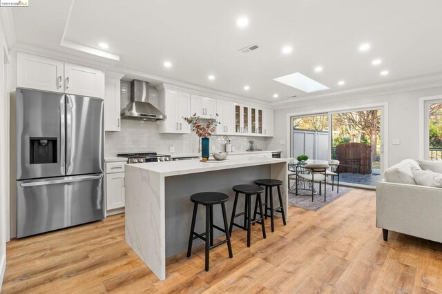 kitchen featuring white cabinets, wall chimney range hood, a skylight, a kitchen breakfast bar, and stainless steel fridge with ice dispenser