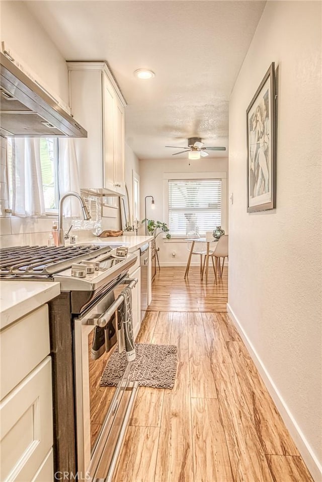 kitchen with ceiling fan, appliances with stainless steel finishes, light wood-type flooring, wall chimney exhaust hood, and white cabinets