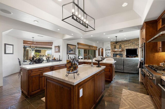 kitchen featuring light stone counters, a center island with sink, a stone fireplace, and decorative light fixtures