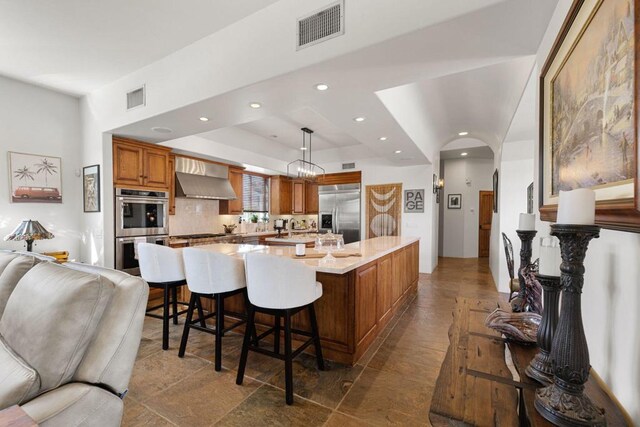 kitchen with stainless steel appliances, a raised ceiling, a spacious island, hanging light fixtures, and wall chimney exhaust hood