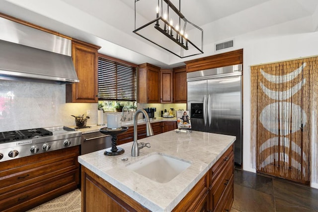 kitchen featuring wall chimney range hood, stainless steel appliances, an inviting chandelier, an island with sink, and sink