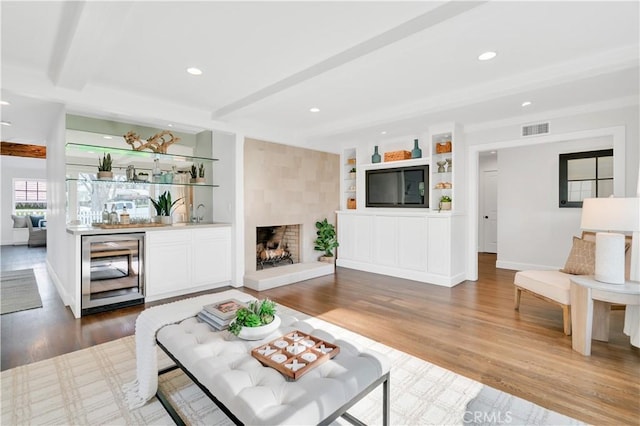 living room with wood-type flooring, beamed ceiling, wine cooler, a large fireplace, and built in features