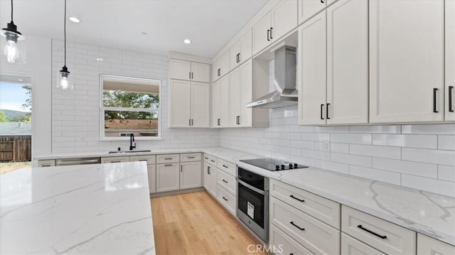 kitchen with wall chimney range hood, oven, sink, white cabinetry, and hanging light fixtures