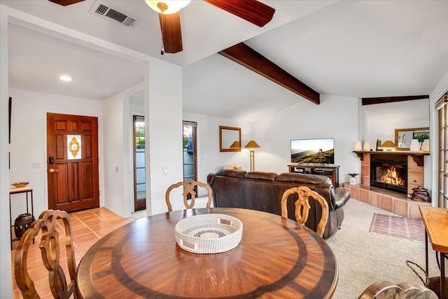 dining room featuring ceiling fan, light colored carpet, a tiled fireplace, and lofted ceiling with beams