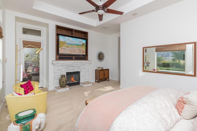 carpeted bedroom with ceiling fan, a tray ceiling, and a stone fireplace