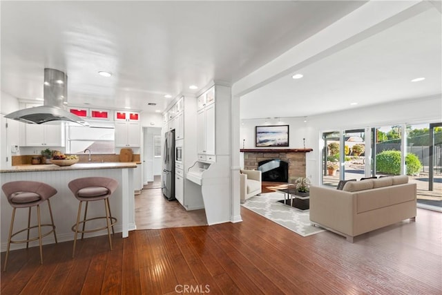 kitchen featuring a fireplace, hardwood / wood-style floors, white cabinetry, stainless steel fridge, and island range hood