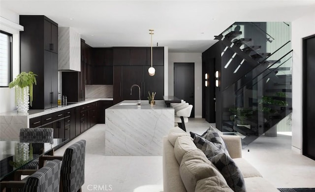 kitchen with dark brown cabinetry, wall chimney exhaust hood, decorative light fixtures, sink, and a center island with sink