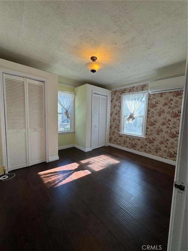 unfurnished bedroom with an AC wall unit, dark wood-type flooring, multiple windows, and two closets