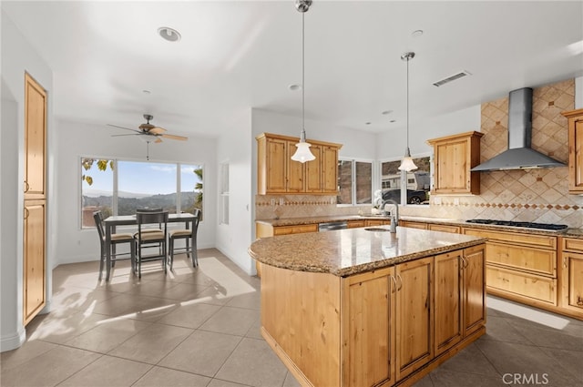 kitchen featuring wall chimney exhaust hood, backsplash, hanging light fixtures, light tile patterned floors, and a center island with sink