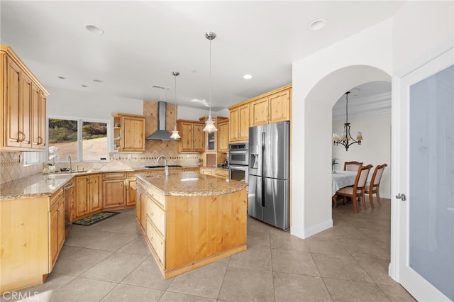 kitchen with decorative light fixtures, wall chimney range hood, appliances with stainless steel finishes, and a kitchen island with sink