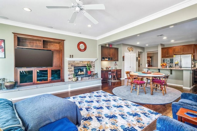 living room with ceiling fan, dark wood-type flooring, ornamental molding, and a stone fireplace