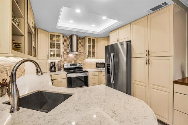 kitchen featuring appliances with stainless steel finishes, a tray ceiling, light stone countertops, wall chimney exhaust hood, and sink