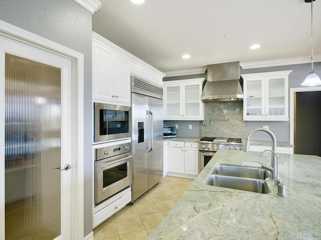 kitchen with wall chimney range hood, pendant lighting, sink, built in appliances, and white cabinets