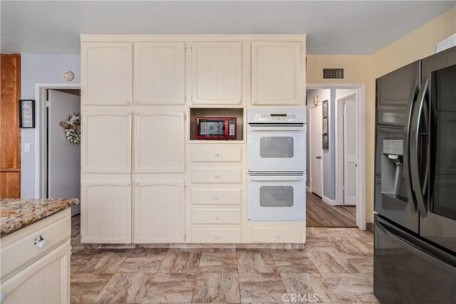 kitchen with black refrigerator with ice dispenser, white double oven, cream cabinetry, and light stone counters