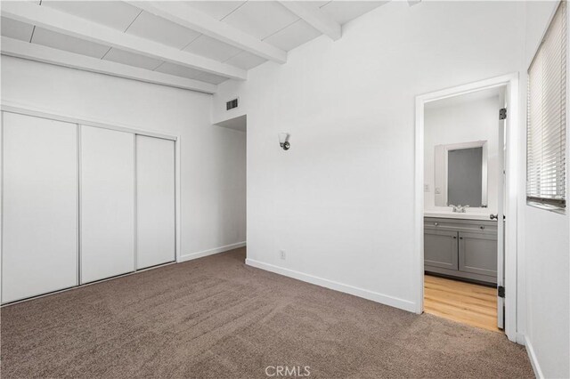 unfurnished bedroom featuring ensuite bathroom, light colored carpet, a closet, and beamed ceiling