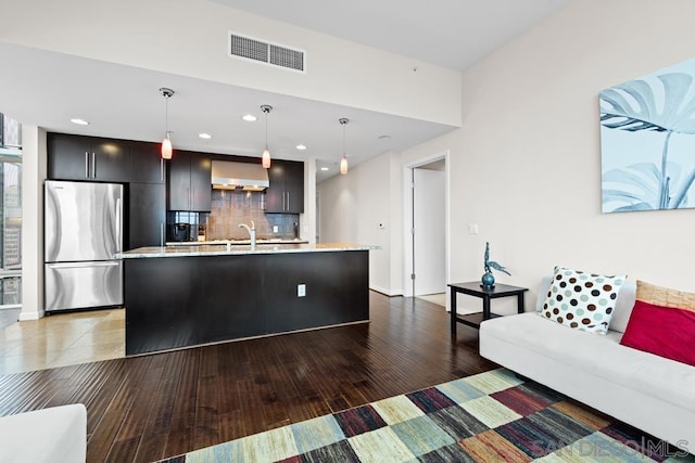 kitchen featuring pendant lighting, stainless steel fridge, a kitchen island with sink, and wall chimney exhaust hood