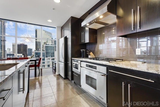 kitchen featuring floor to ceiling windows, appliances with stainless steel finishes, backsplash, wall chimney range hood, and light stone counters