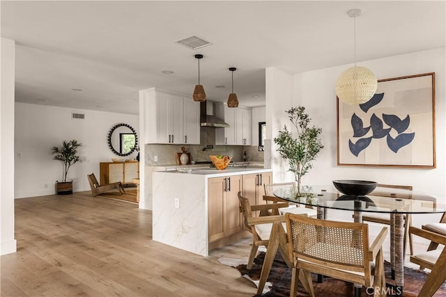 kitchen with wall chimney exhaust hood, white cabinetry, backsplash, hanging light fixtures, and light wood-type flooring