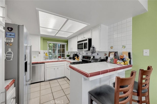 kitchen featuring tile countertops, kitchen peninsula, decorative backsplash, appliances with stainless steel finishes, and white cabinets