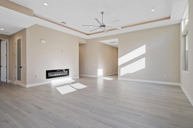 unfurnished living room with ceiling fan, a tray ceiling, ornamental molding, and light hardwood / wood-style flooring