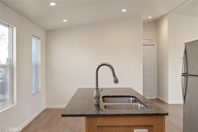 kitchen featuring an island with sink, dark stone countertops, sink, stainless steel refrigerator, and light hardwood / wood-style flooring