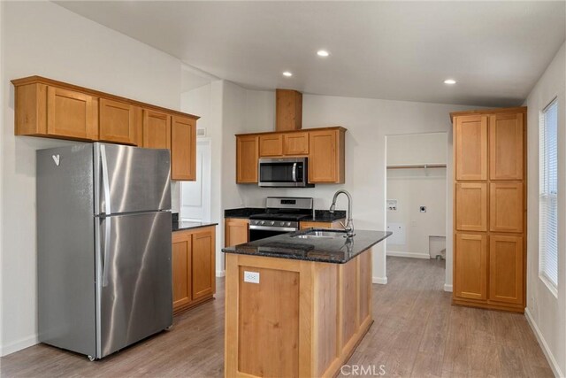 kitchen with lofted ceiling, stainless steel appliances, dark stone countertops, light hardwood / wood-style flooring, and a center island with sink