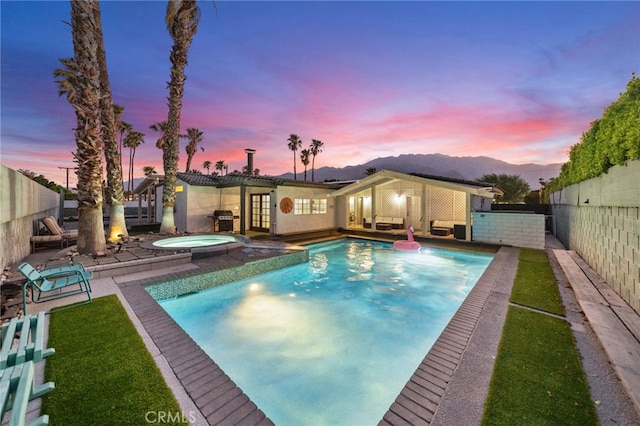 pool at dusk with a patio area, a mountain view, grilling area, and an in ground hot tub