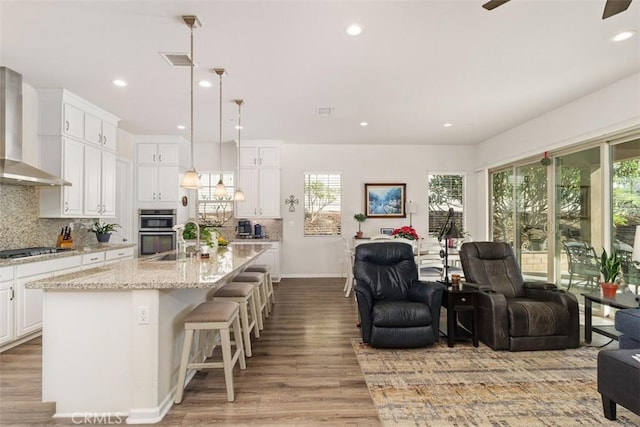 kitchen featuring wall chimney exhaust hood, stainless steel appliances, an island with sink, pendant lighting, and white cabinets