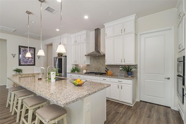 kitchen featuring stainless steel fridge, white cabinetry, an island with sink, and wall chimney range hood