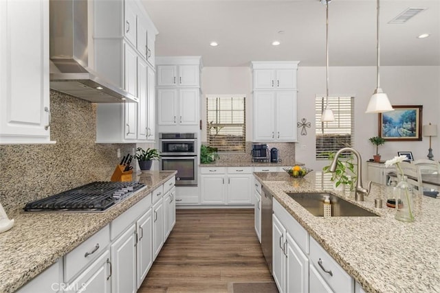 kitchen with appliances with stainless steel finishes, sink, wall chimney range hood, pendant lighting, and white cabinetry