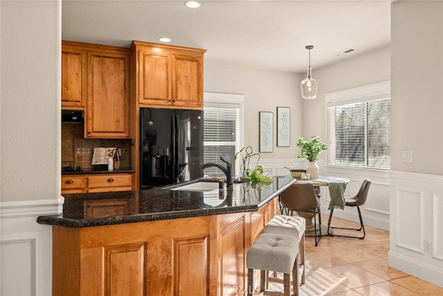 kitchen with black refrigerator with ice dispenser, dark stone counters, sink, hanging light fixtures, and light tile patterned floors