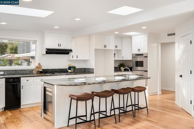 kitchen with dark stone countertops, white cabinetry, a skylight, appliances with stainless steel finishes, and an island with sink