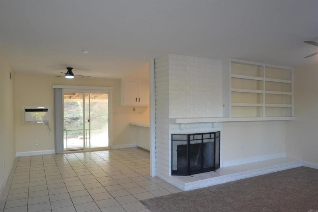 unfurnished living room featuring light tile patterned flooring, built in shelves, a wall mounted AC, a brick fireplace, and ceiling fan