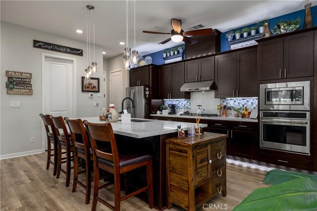 kitchen with a center island with sink, decorative backsplash, hanging light fixtures, appliances with stainless steel finishes, and dark brown cabinets