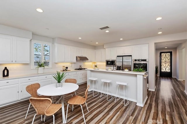 kitchen with appliances with stainless steel finishes, dark hardwood / wood-style flooring, white cabinets, and a kitchen island with sink