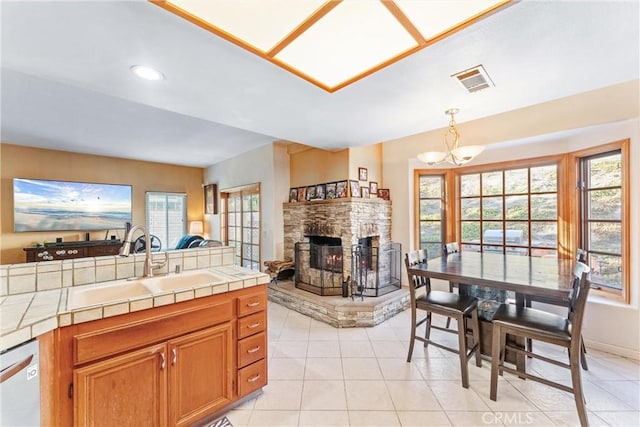 kitchen with a stone fireplace, tile counters, hanging light fixtures, dishwashing machine, and sink