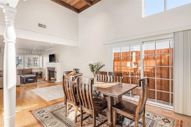 dining space with a brick fireplace, hardwood / wood-style flooring, a towering ceiling, beam ceiling, and decorative columns