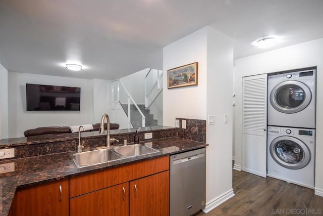 kitchen featuring dark wood-type flooring, dark stone counters, stacked washing maching and dryer, stainless steel dishwasher, and sink