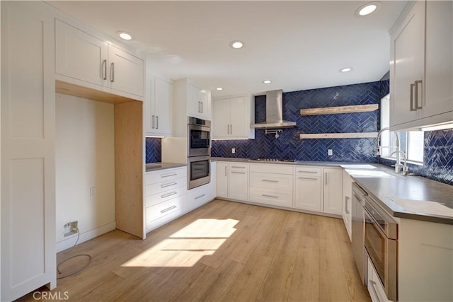 kitchen with appliances with stainless steel finishes, decorative backsplash, white cabinets, wall chimney range hood, and light hardwood / wood-style flooring