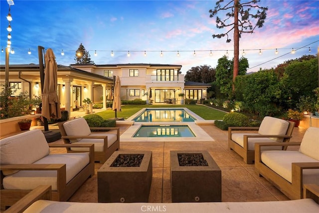 back house at dusk with a pool with hot tub, an outdoor living space with a fire pit, a patio area, and a balcony