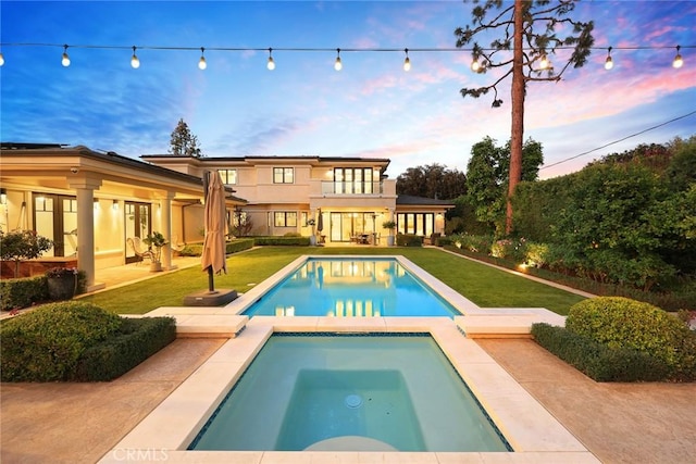 back house at dusk featuring a pool with hot tub, a yard, a balcony, and a patio